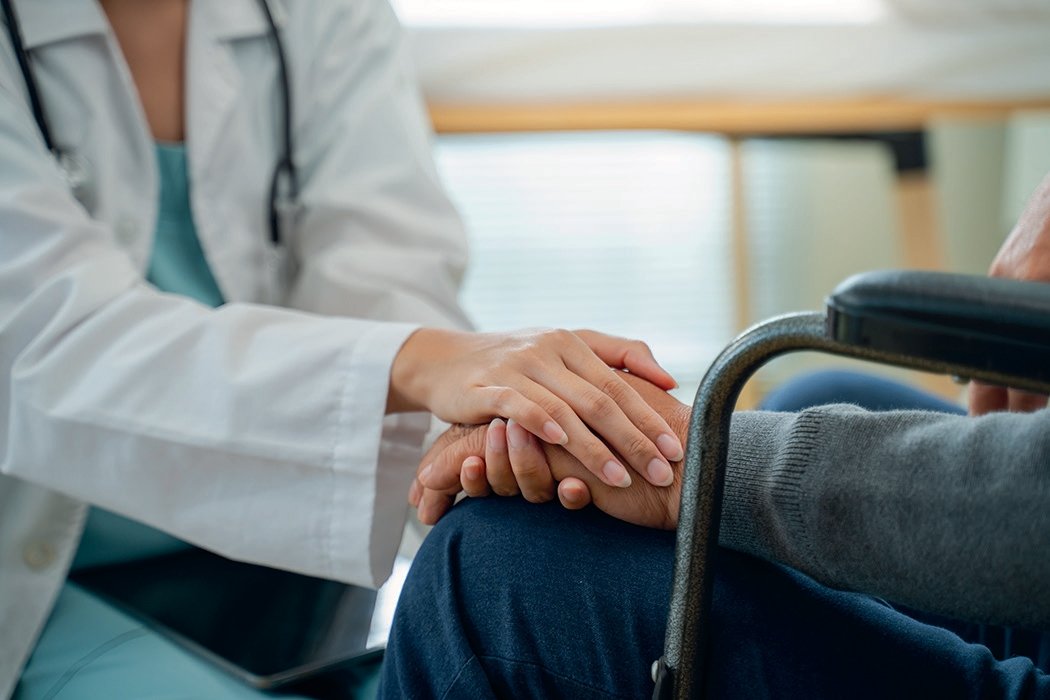 Médica de bata branca segurando carinhosamente a mão de idoso em cadeira de rodas numa residência sénior